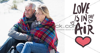 Composite image of happy couple wrapped up in blanket sitting on the beach