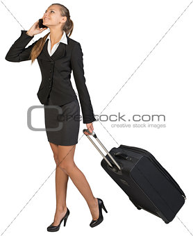 Businesswoman walking with wheeled suitcase, talking on the phone, smiling