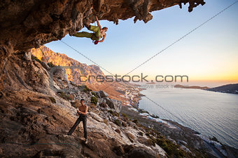 Male rock climber climbing on a roof in a cave, his partner belaying