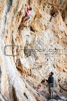 Young woman lead climbing on natural cliff, belayer watching her