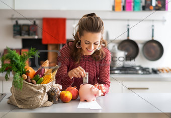 Portrait of happy young housewife putting money into piggy bank 