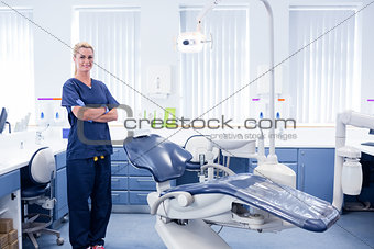 Dentist in blue scrubs standing with arms crossed beside chair