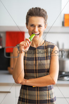 Portrait of happy young housewife eating cucumber in kitchen