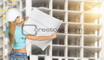 Woman in helmet standing backwards and holding paper sheet. Building under construction as backdrop
