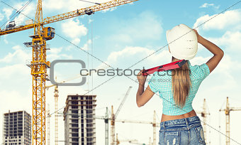 Woman in helmet stands backwards, holding building level. Looks at crane. Construction site as backdrop