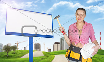 Woman holding hammer and helmet. Green hills, road, empty billboard, buildings as backdrop