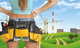 Woman in tool belt stands back. Green hills and cranes with heat power stations on background