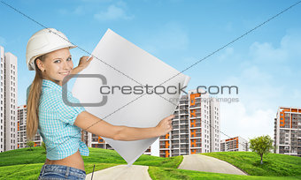 Woman in helmet holds white paper. Looking at camera, smiling. Green hills with road and buildings