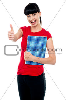 Pretty teenager holding notebook and gesturing thumbs up