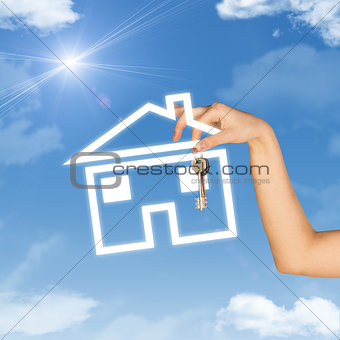 Hand holding house icon and key. Background of sky, clouds, sun