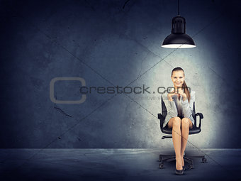 Woman wearing jacket, blouse sitting with legs crossed. Background concrete wall