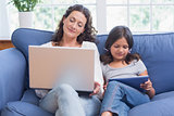 Happy mother and daughter sitting on the couch while using laptop and tablet