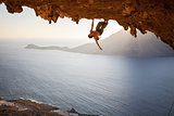 Female rock climber posing while climbing along roof in cave at sunset