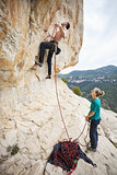 Rock climber going to clip rope at beginning of route, girl belayer watching him