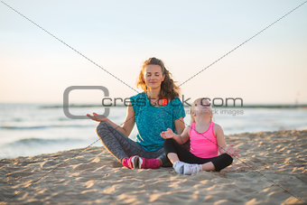 Healthy mother and baby girl doing yoga on beach in the evening