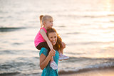 Happy baby girl sitting on shoulders of mother on beach in the e