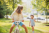 Mother playfully riding a blonde girl's bike in a sunny park