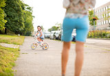 Smiling girl rides her bike on city sidewalk as mother watches
