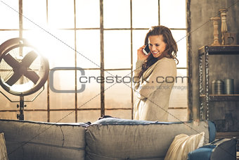 Smiling brunette in loft stands talking on the phone behind sofa