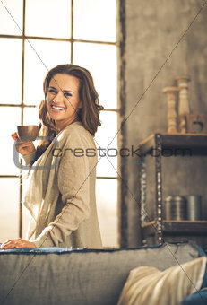 Brunette holding cup of coffee while smiling in loft apartment