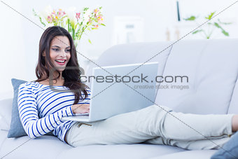 Smiling beautiful brunette relaxing on the couch and using her laptop