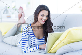 Smiling beautiful brunette relaxing on the couch and using her laptop