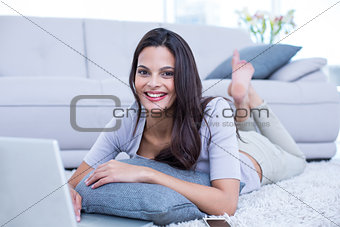 Smiling beautiful brunette lying on the floor and using her laptop