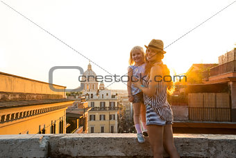 Mother holding daughter standing on ledge at sunset in Rome