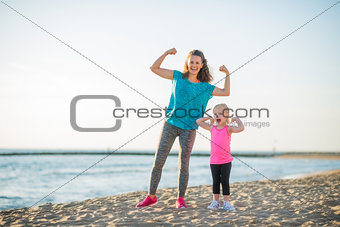 Happy mother and daughter in fitness gear on beach flexing arms