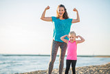 Joyful mother and daughter in fitness gear on beach flexing arms