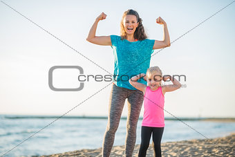 Joyful mother and daughter in fitness gear on beach flexing arms