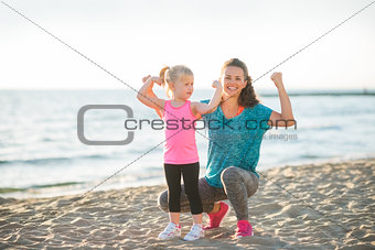 Young mother and daughter in fitness gear on beach flexing arms