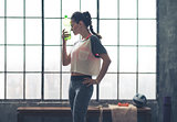 Woman by window in loft gym holding water bottle to head