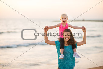 Joyful mother holding daughter on shoulders on beach at sunset