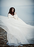 Beautiful sad young woman in white dress standing on sea beach