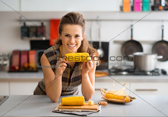 Smiling woman leaning on kitchen counter holding corncob