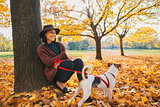 Young woman with cute dog sitting under tree in autumn park