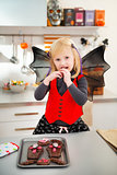 Blond girl in bat costume eating Halloween biscuits in kitchen