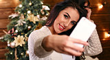 people, holidays and technology concept - beautiful brunette woman in white  sweater taking selfie picture by smartphone over christmas tree and living room background
