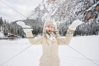 Happy woman in fur hat throwing up snow while standing outdoos