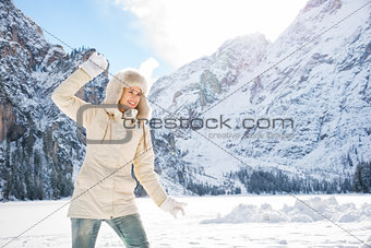 Happy woman in coat and fur hat throwing snow ball outdoors