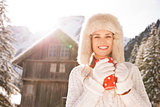 Young woman with red cup standing near cosy mountain house