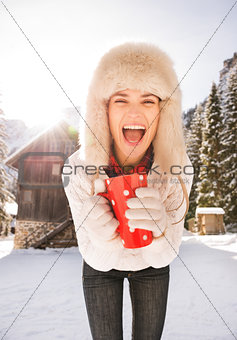Cheerful woman with red cup standing near cosy mountain house