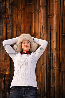 Relaxed woman in knitted sweater and furry hat near wood wall
