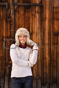Woman in white knitted sweater and furry hat near wood wall