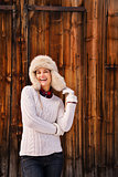 Happy woman in white sweater and furry hat near rustic wood wall