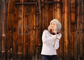 Happy woman in furry hat near rustic wood wall looking up