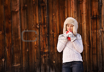 Pensive young woman in furry hat with cup near rustic wood wall