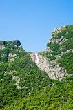 Steep cliffs covered in trees with distant waterfall under blue 