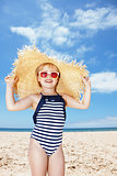 Happy girl in striped swimsuit and big straw hat on white beach